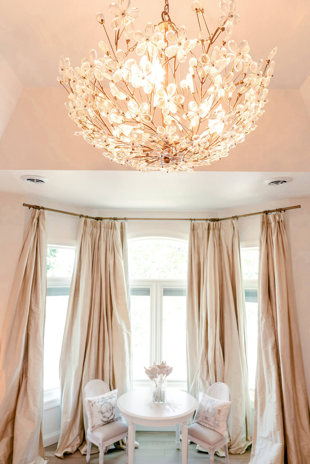 Storybook-38 Elegant bedroom with a sculptural floral chandelier, tall windows, and softly draped neutral curtains