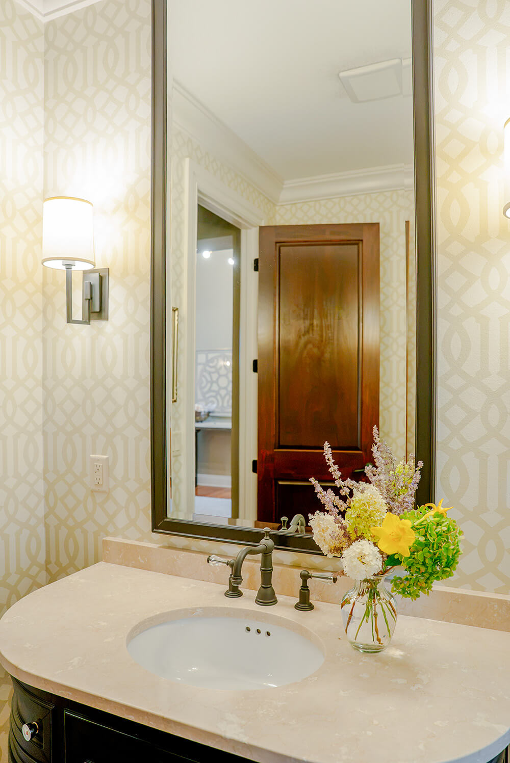 taylor-jane-project-1 Elegant bathroom vanity with a marble countertop, dark wood cabinetry, patterned wallpaper, large framed mirror, and fresh floral arrangement.