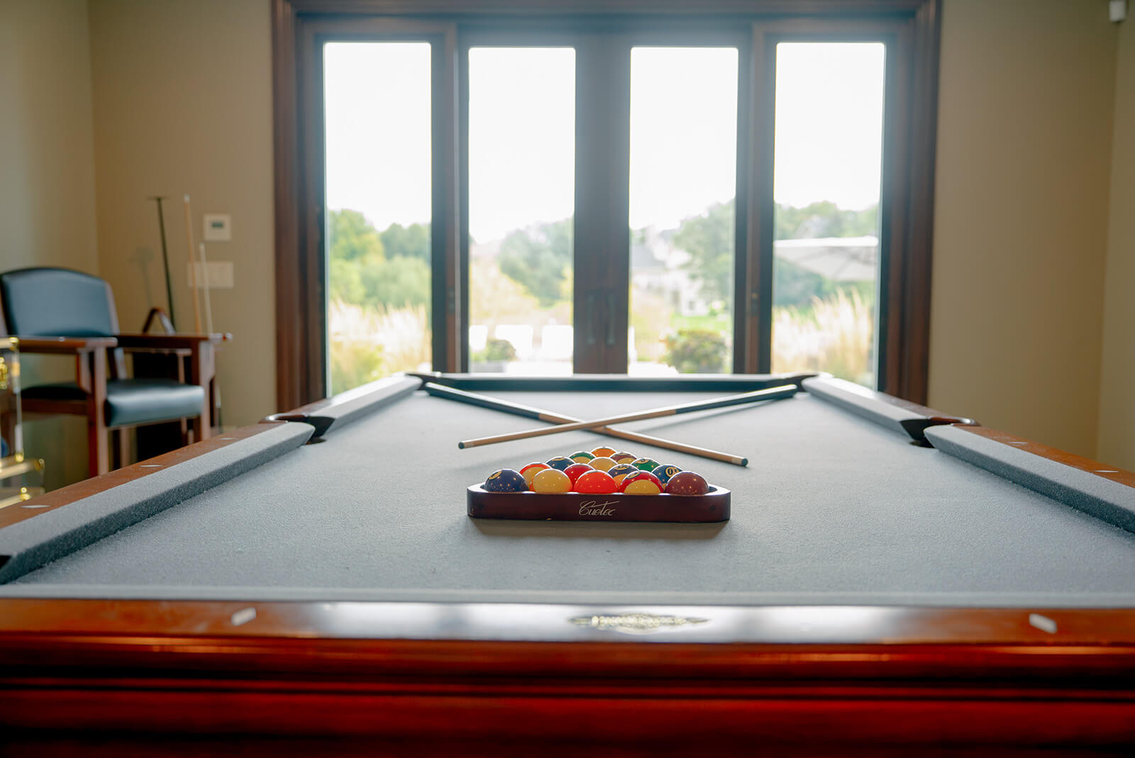taylor-jane-project-17 Luxury game room with a billiards table, crossed pool cues, and a rack of colorful billiard balls in front of large windows.